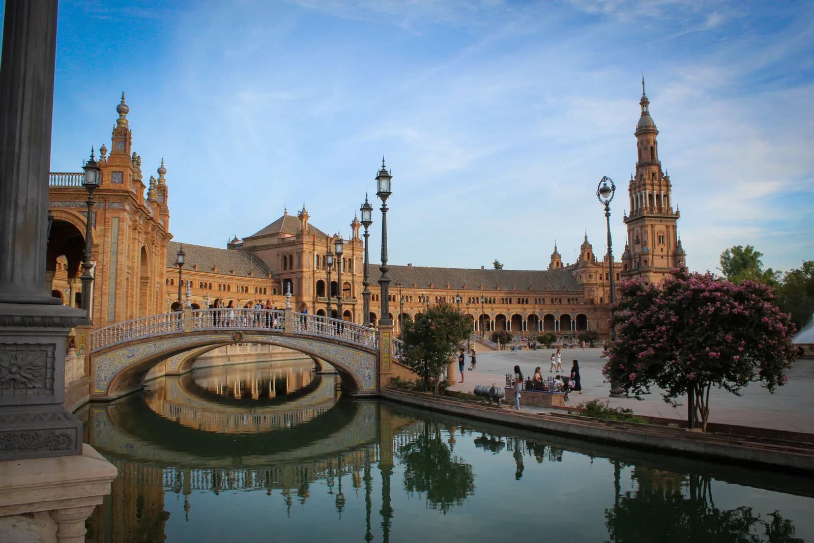 Plaza de España w Sewilli: mostek z ceramiczną balustradą nad kanałem, półkole ceglanych budynków i wieża w wieczornym świetle