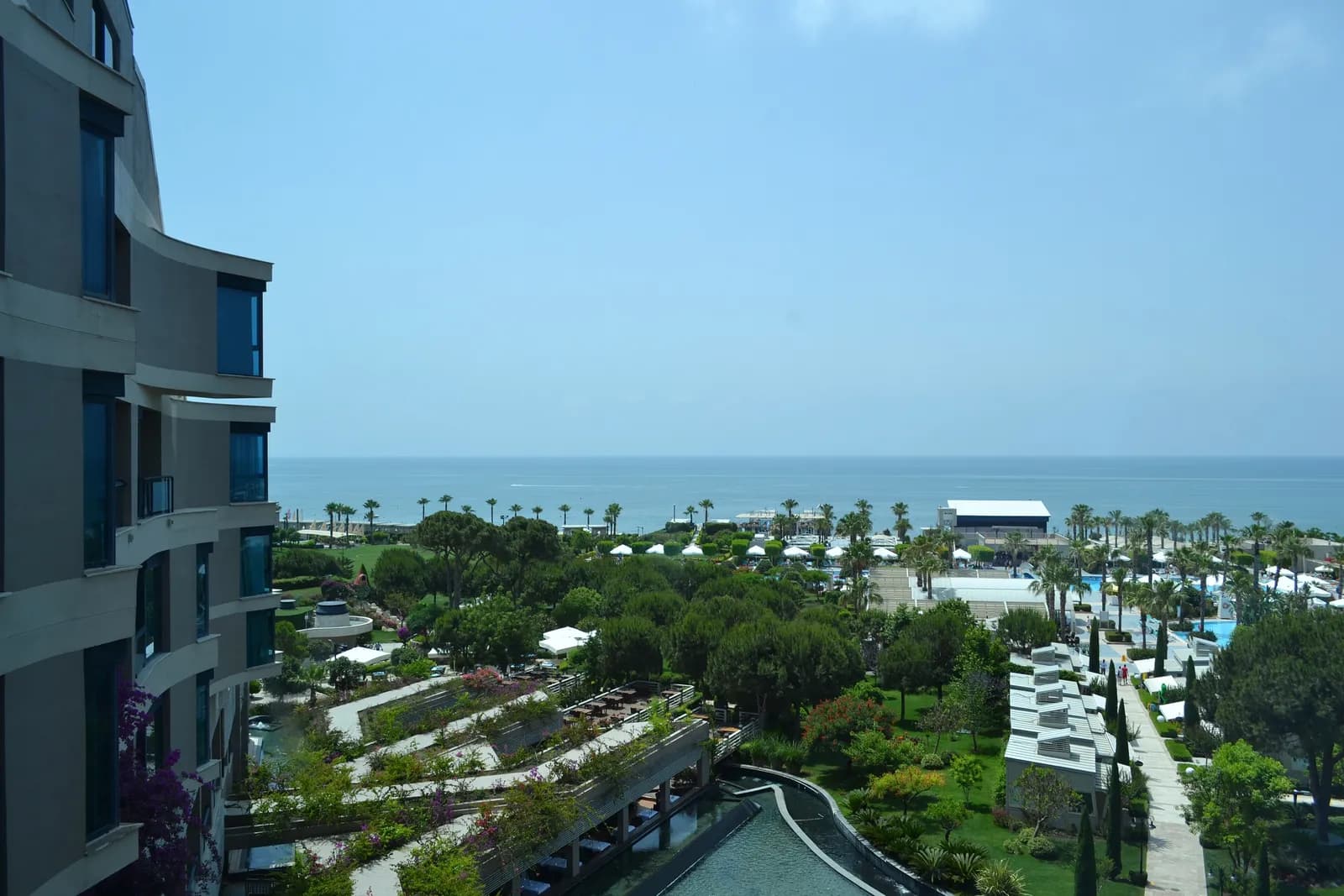 Widok z wyższego piętra hotelu Susesi na tarasowe ogrody, niskie białe pawilony i baseny, w tle linia palm i morze