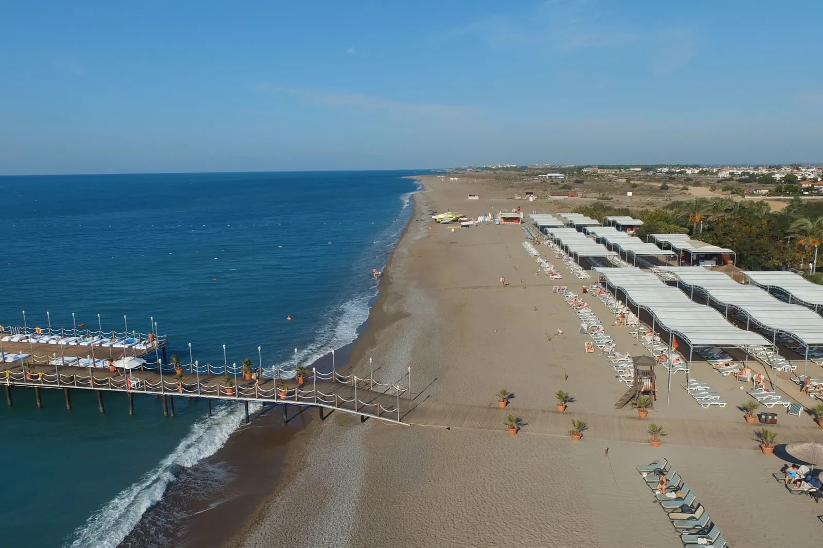 Długa piaszczysta plaża z pomostem i rzędami leżaków pod zadaszeniami, widok z góry