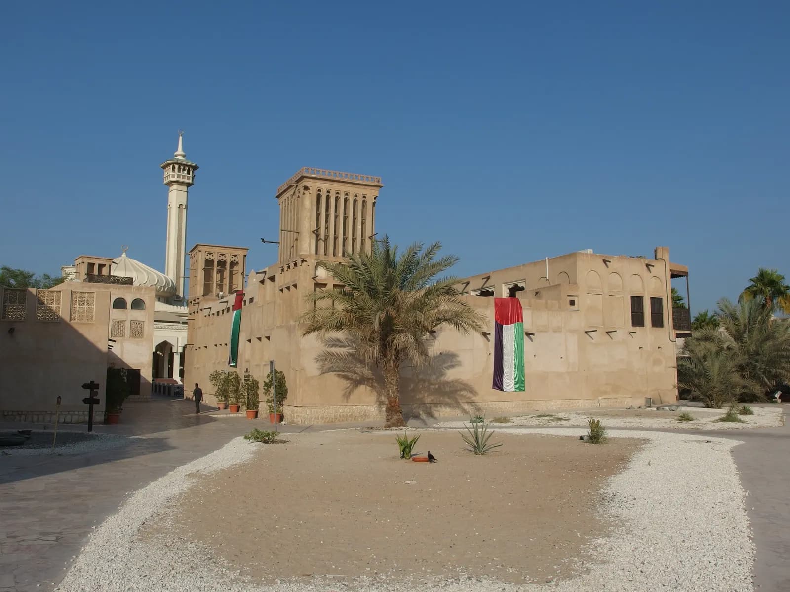 Tradycyjne domy z wieżami wiatrowymi w historycznej dzielnicy Al Fahidi (Al Bastakiya) w Dubaju, na murach flagi Emiratów, w tle minaret